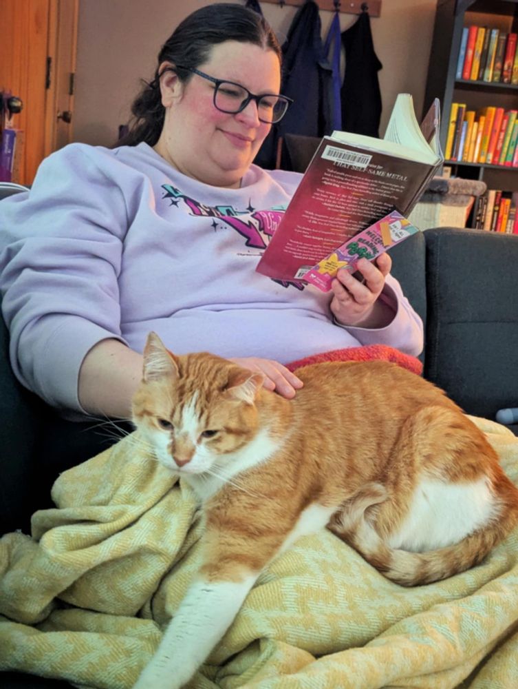 Jill reading a book and petting big boi orange and white cat stretched on her lap. Photo taken by her husband. Not pictured.