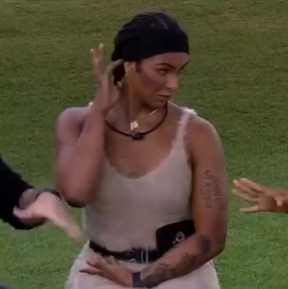 a woman in a white tank top is standing on a lush green field with her arms crossed .