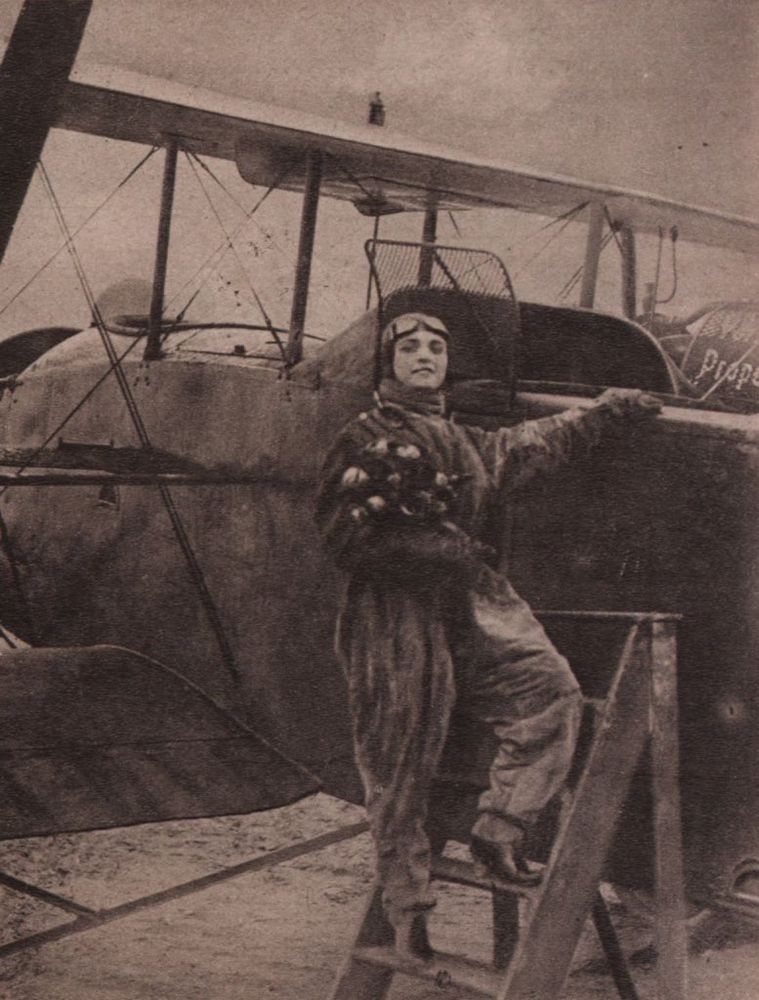 Pola Negri wearing a flight suit, holding a bouquet of flowers and posing while climbing up a ladder to board the plane she‘s standing in front of.