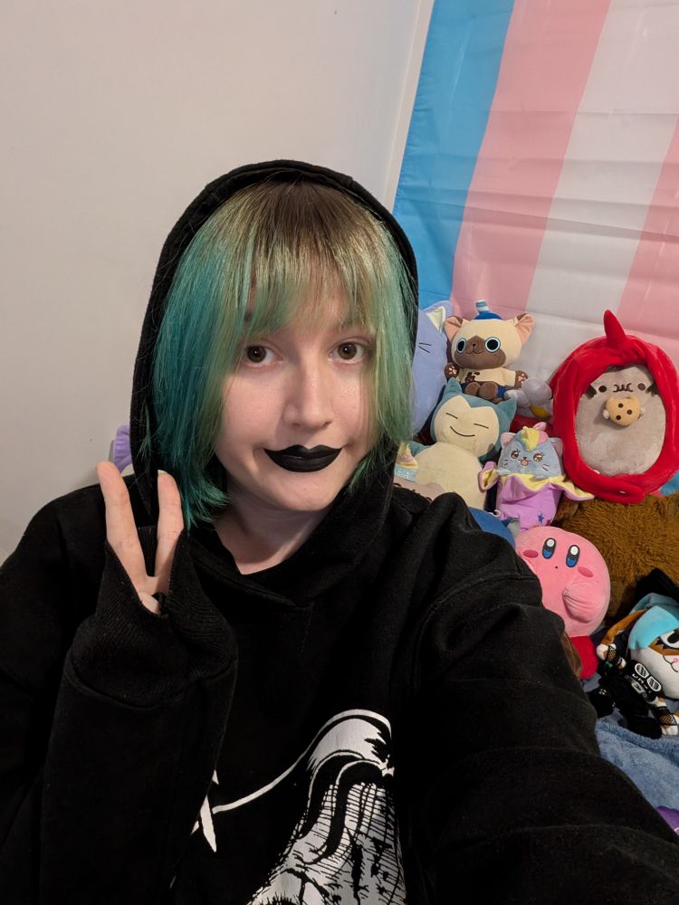 Revy wearing a hoodie
It's slightly on her head
She is wearing black lipstick.
Many plushies are in the background, Kirby, bunlith, Pusheen, snorlax & others