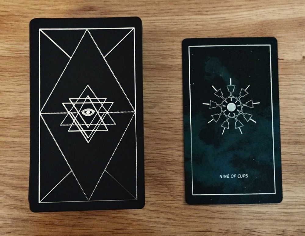 The Nine of Cups for the Synesthesia Tarot deck. The deck is black with bright silver lines creating a geometric design of triangles with a simple drawing of an eye in the middle. The card is nine cups arranged in a circle around a simple drawing of a sun with rays. Each cup has a small line pointing from the middle of the goblet. The background is blue smoke, and there is a thin, bright silver border. 