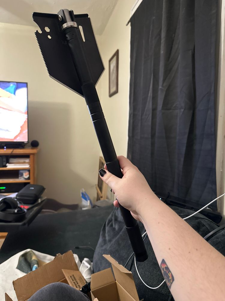 Photo showing an arm holding a 28” long shovel. The shovel has a serrated edge as well as various other utility-focused cutouts. The end of the shovel is folded inwards to reduce its overall size. 