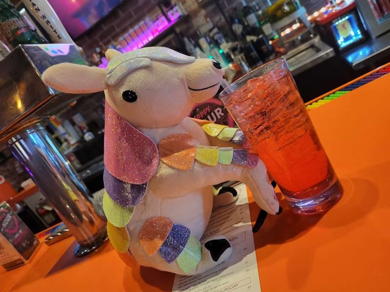 A plush armadillo with various sparkly colored "plates" running down its back, legs, and arms, "sipping" a drink out of a straw at a bar.