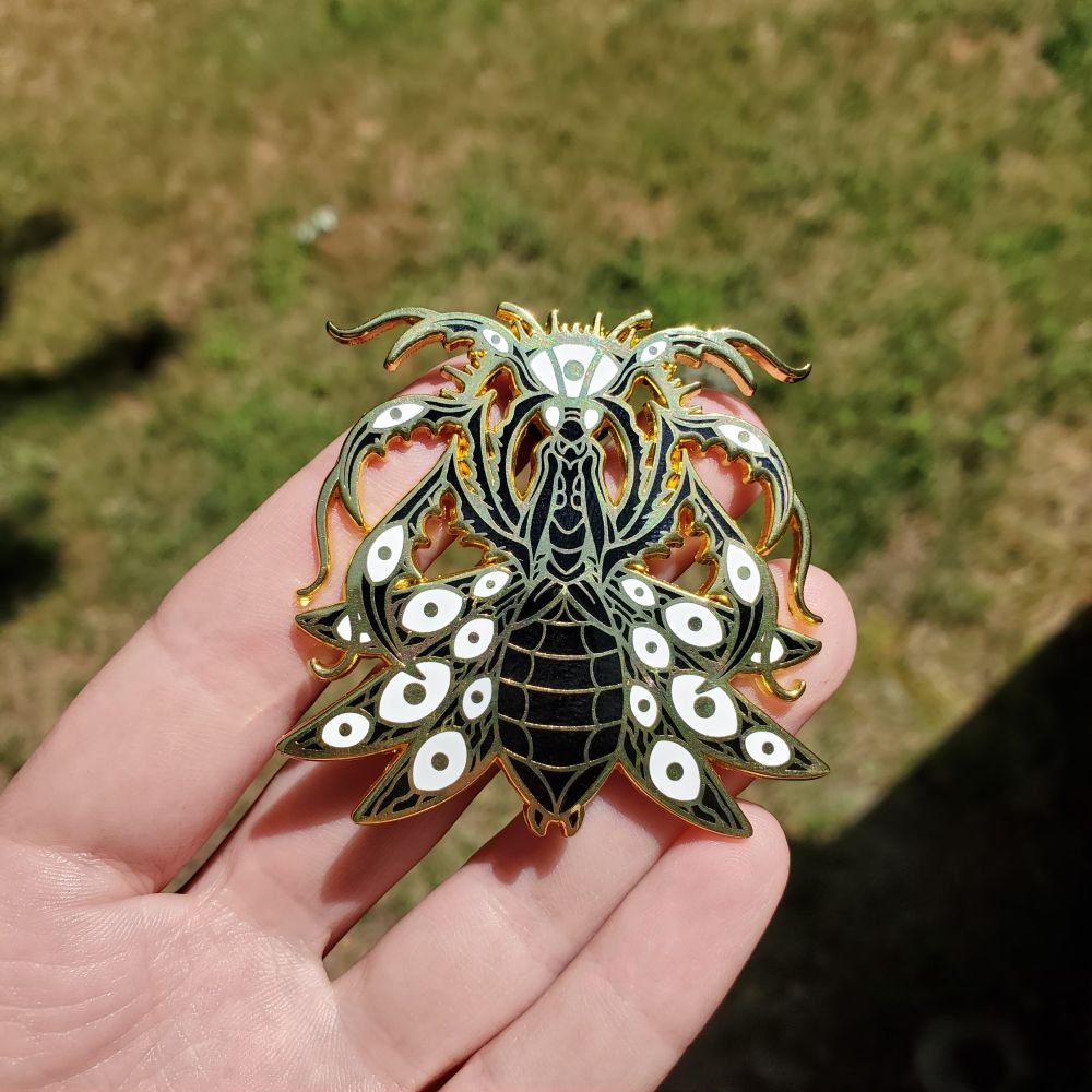 A hand holding a golden, black, and white multi limbed praying mantis pin with eyes all over it.
