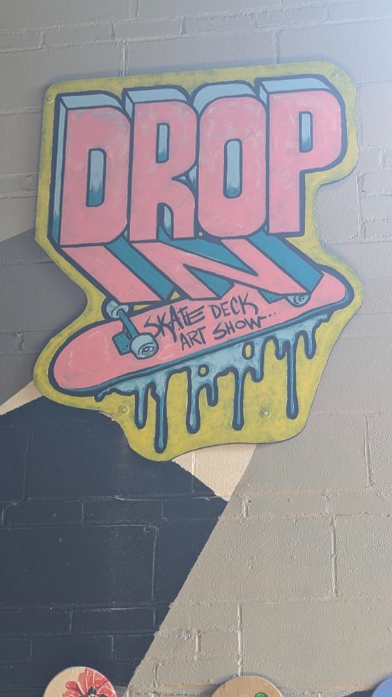 Photograph of a wooden graffiti style sign that reads "drop in skate deck art show" hung on a wall.