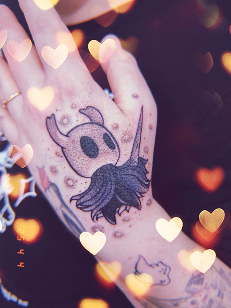 the night from hollow knight tattooed on Mae's hand. their cloak is billowing, their nail raised, and there are twinkling lights around them.