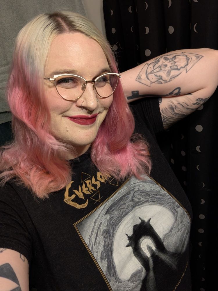 Mae with her ombre blonde into pink hair down, her left hand behind her head so her elbow is pointing out and

she is wearing white and gold glasses and dark red lipstick, as well as an Everson Poe shirt