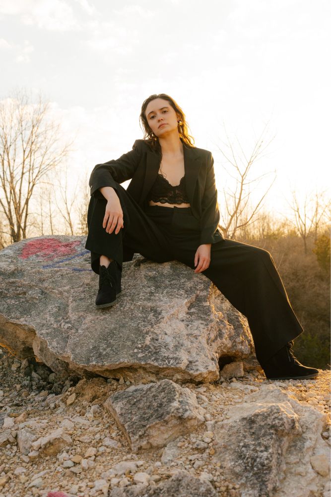 Actress & Model Brandi Botkin wearing a wide leg pantsuit, blazer unbuttoned and open showing a black lace bralette. She sits on top of a jagged boulder with the sun behind her. 