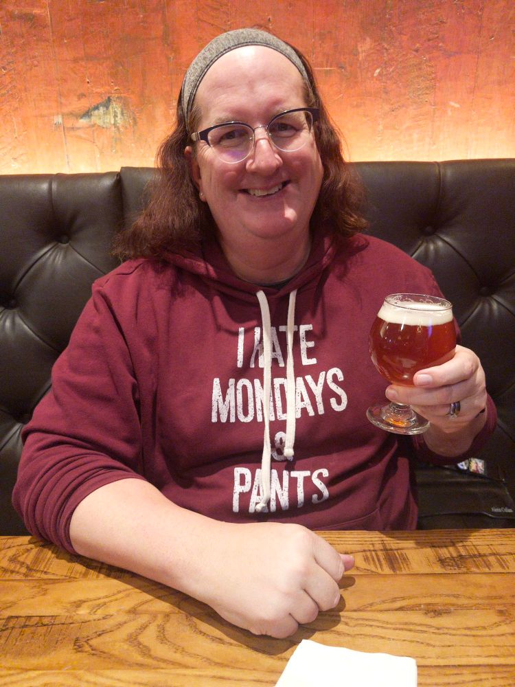 Jamie is sitting in a restaurant booth, raising a glass in toast. They are smiling and wearing glasses. They are also wearing a maroon hoody with a phase in white letters across the front. It reads “I hate Mondays and pants.”