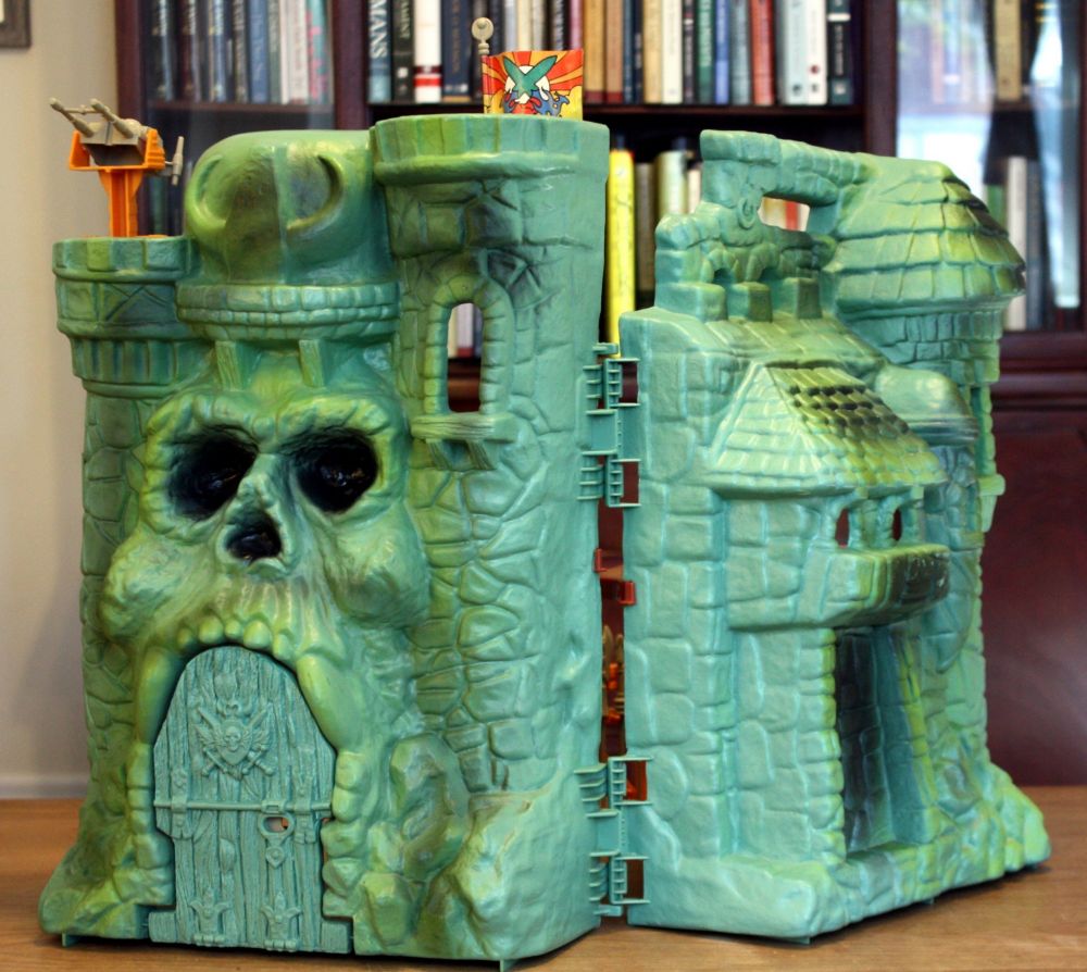Cast of Grayskull toy. Plastic toy castle. It is green in color, has an engraved pattern that looks like stone, and an entrance that is shaped like a skull face with a drawbridge for a mouth.