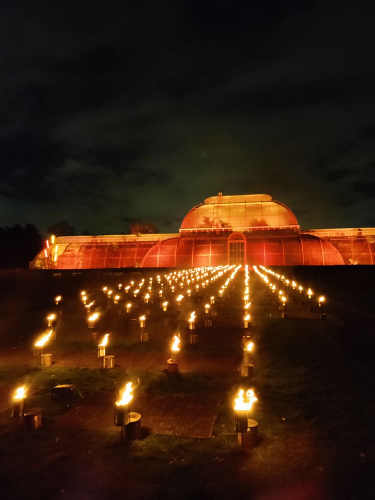A large glasshouse lit orange surrounded by torches on the grass.