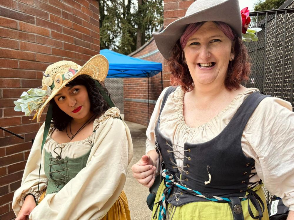 Two women in Renaissance Faire garb pose for a photo between two brick walls.  The woman on the right is laughing, while the woman on the left looks at her with a mix of affection and bemusement.  
