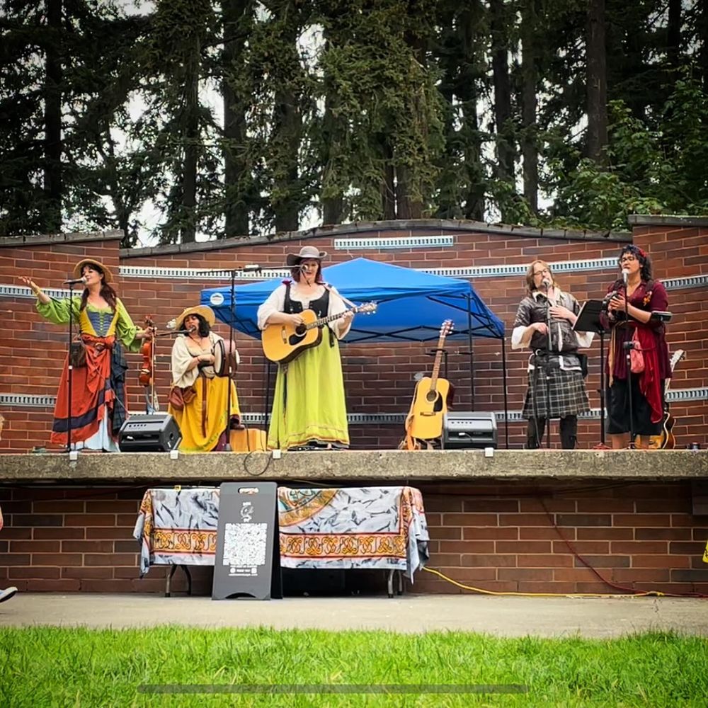 A Celtic Folk band in brightly colored costumes performs on a brick stage with tall trees in the background.  