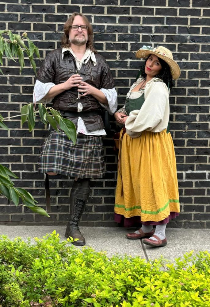 A man and woman in Renaissance Faire garb pose for the camera while leaning against a brick wall.  