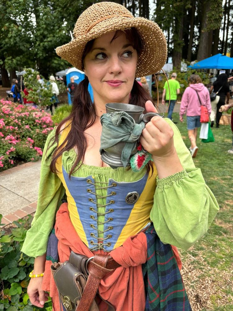 A woman in Renaissance Faire stands in grass with trees and flowers in the background.  She holds a pewter tankard and gazes up and to the left with a wry smile.  