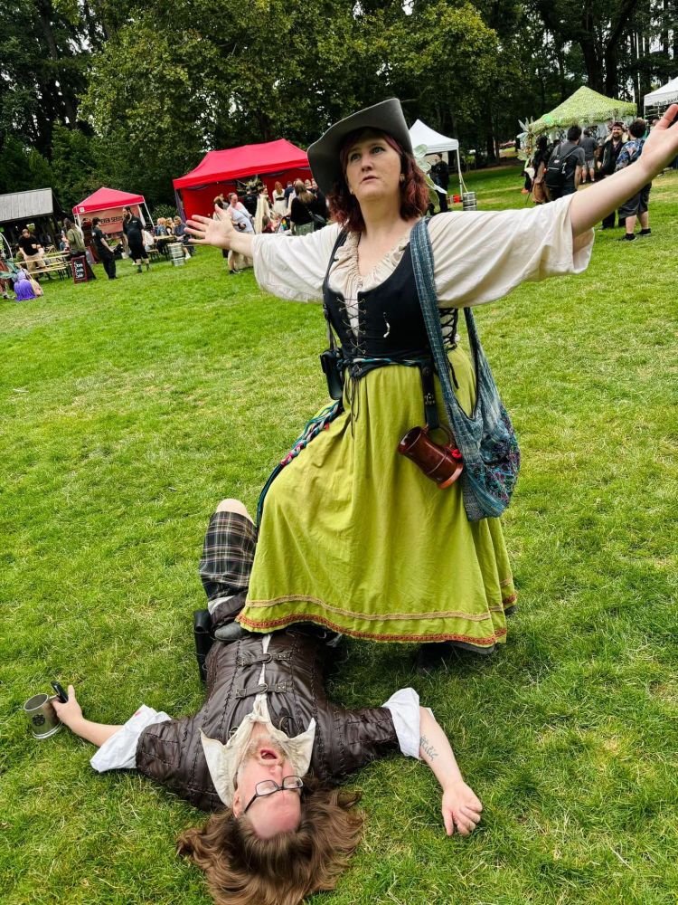 A woman and man in Renaissance Faire garb pose in a joking manner, with the man on the ground pretending to be in pain, while the woman's foot is on his chest and her arms are spread out victoriously.  It looks like she has defeated him in battle.  