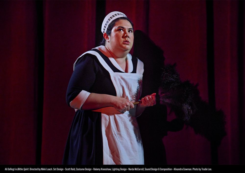 Against a red curtain, a woman in a maid’s outfit tightly holds a feather duster while looking around nervously. 
