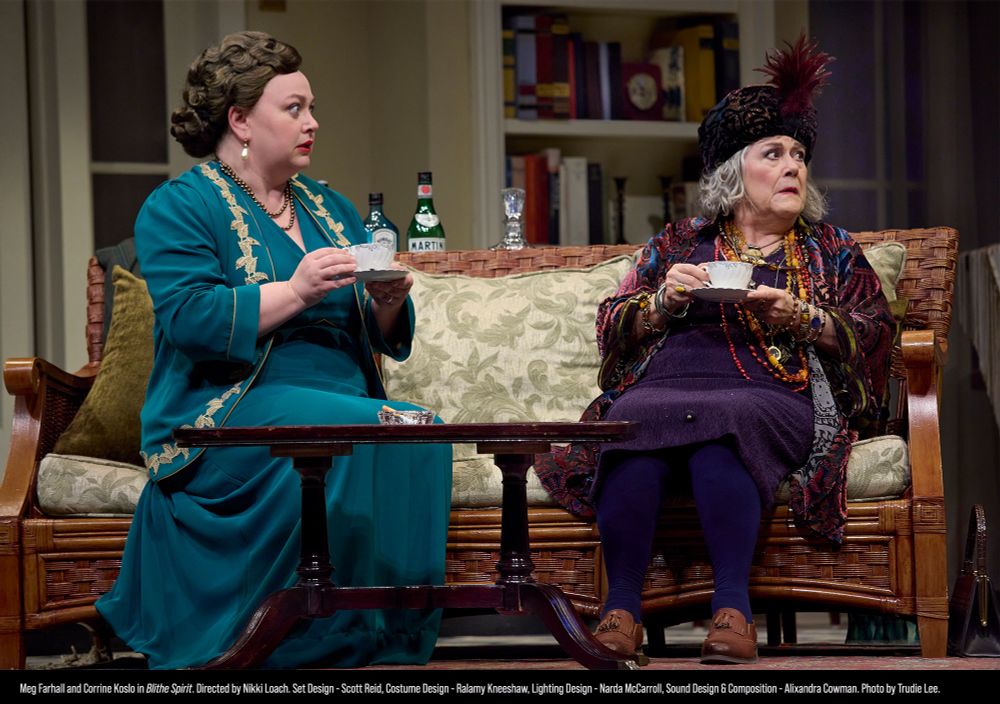 A woman in fancy evening wear and a medium, many wearing layers of jewellery, sit on a couch holding tea cups. They look at something out of frame with shocked expressions.