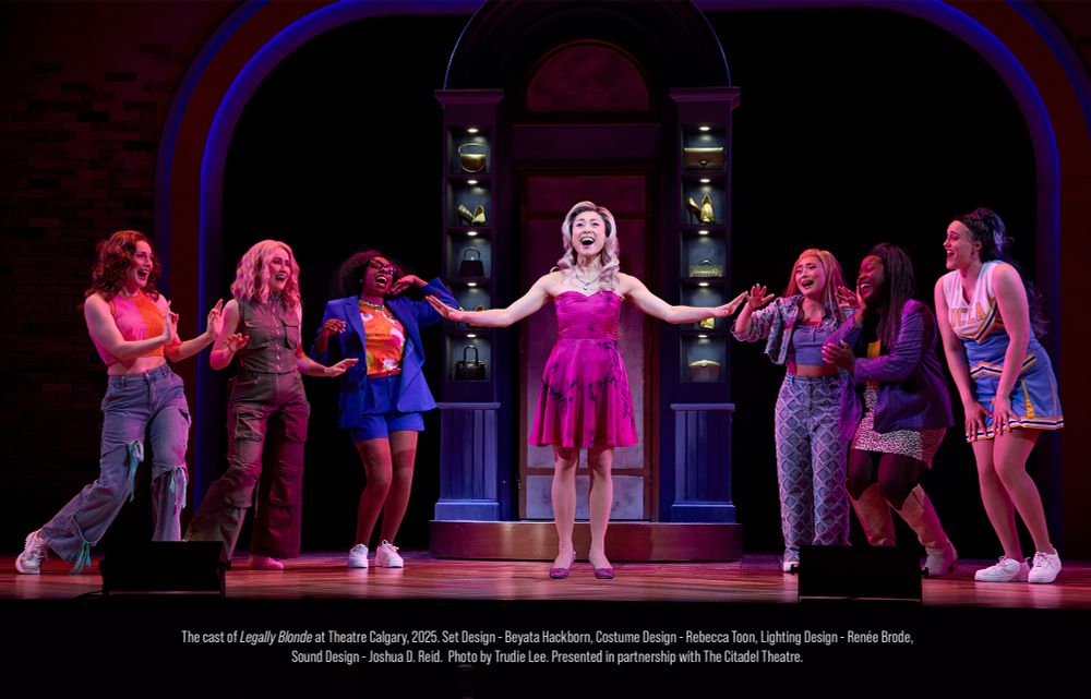 Elle Woods stands centre stage wearing a fuchsia pink dress and shoes, with her arms outstretched, singing and looking happy. She has wavy blonde hair. She is surrounded by her friends, also singing, who look at her adoringly. The background is a fancy dressing room with a shoe display.