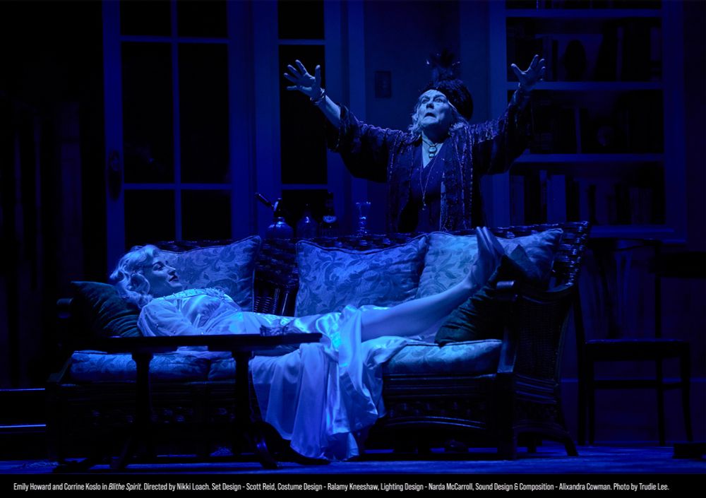 In a dark living room, a woman in a white dress lays on a couch looking amused, as a medium stands over her with arms raised, appearing to do a ritual. 