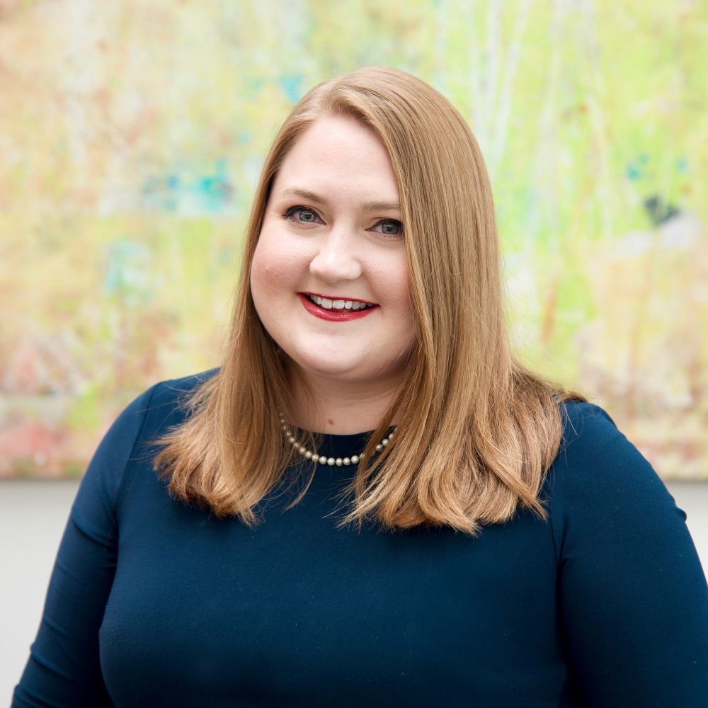 Professional headshot of Jillian wearing a deep green, long sleeve top and pearls. She stands in front of an abstract green painting. 