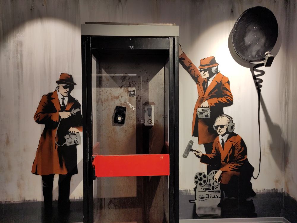 eine schlichte Telefonzelle um die herum drei Agenten mit Mikrofonen und Aufnahmgeräten an die Wand gesprüht sind. sie tragen orange Trechcoats und Hüte, einer trägt einen Kopfhörer, weiße Hemden und schwarze Krawatten