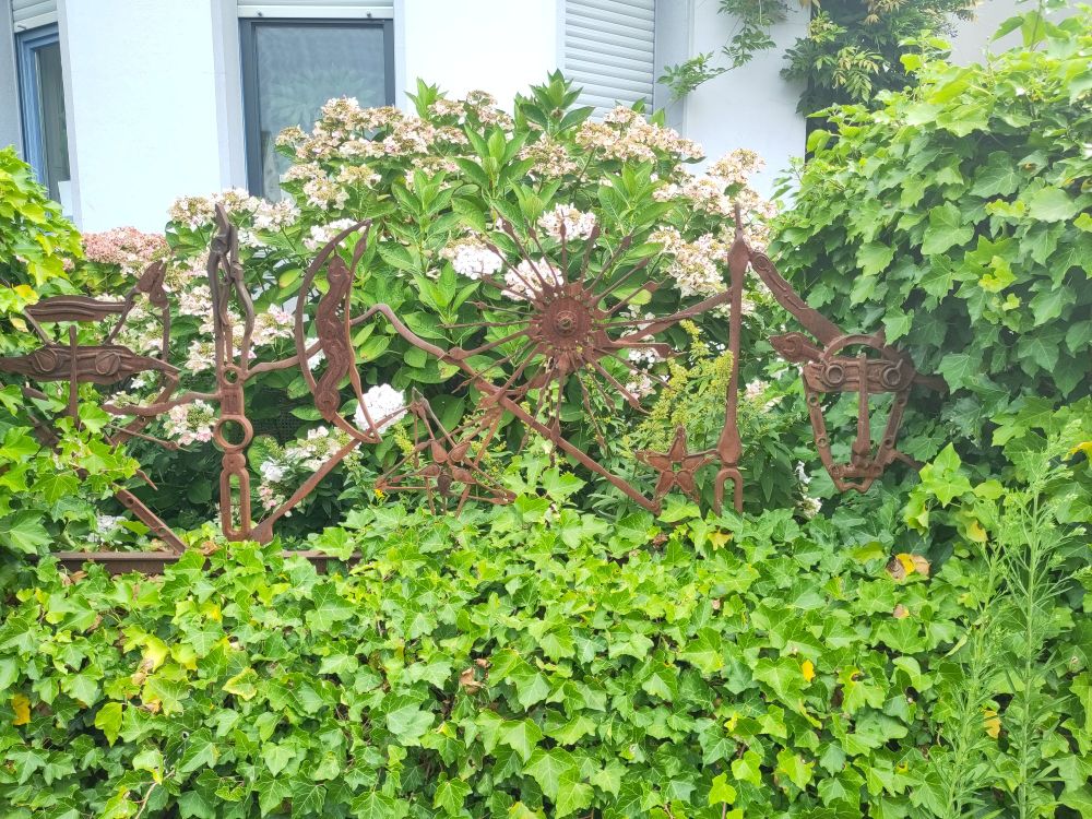 ein Gartenzaun, zusammengeschmiedet aus alten Werkzeugrohlingen. mittig ist ein Mond und eine Sonne zu entdecken. rechts ein Ziegenkopf. links erkenne ich leider nichts. eingerahmt ist der Zaun von einer efeuüberwucherten Mauer