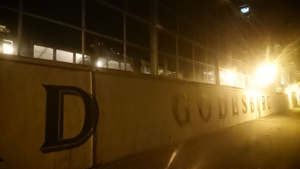 eine weiße Mauer bzw Hauswand. ca 1,50m hoch. darüber, über die komplette Fassade, Glas. von der Beschriftung der Mauer ist ein ‚D‘ von BAD und ‚GODESBERG‘ im Ausschnitt zu sehen. im hinteren Teil des Weges steht eine Laterne die die Nacht erhellt und „ganz GODESBERG“ überbelichtet