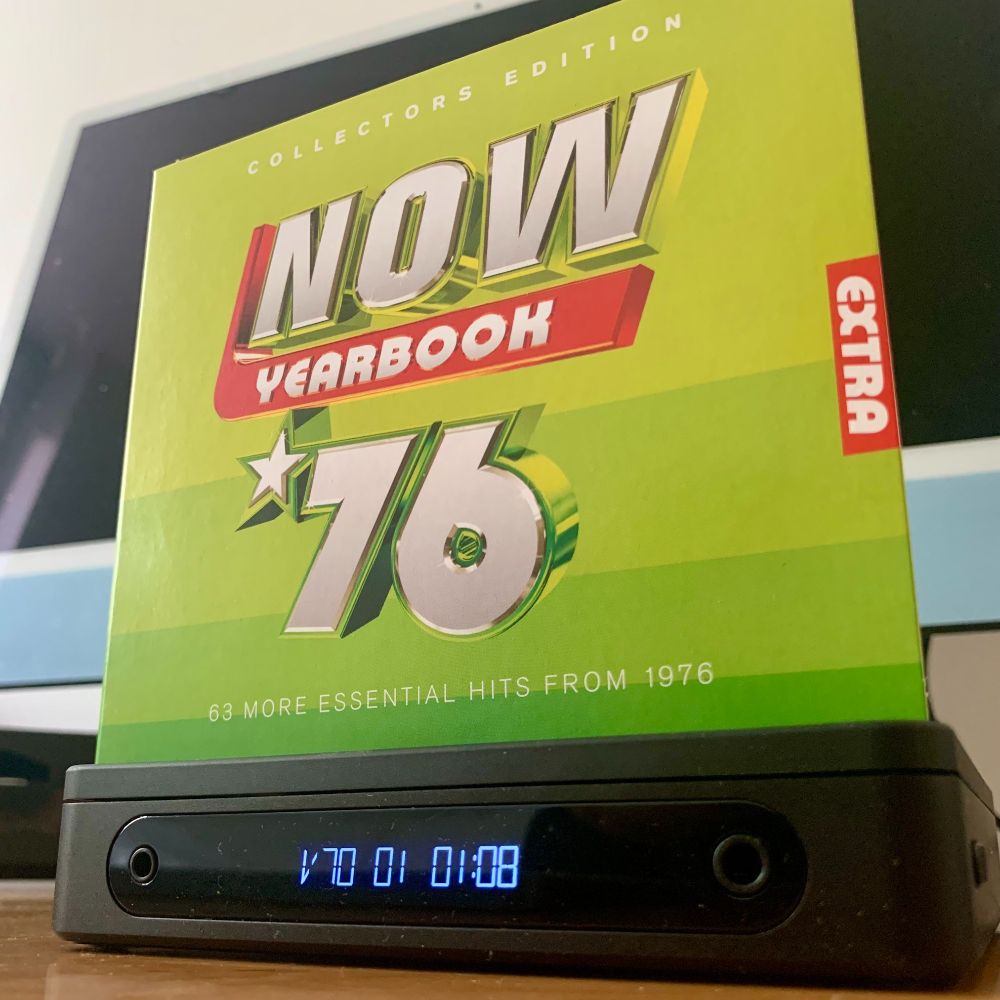 The bright green box of the CD set “Now Yearbook ’76: Collectors Edition” sits on top of a black CD player. The display on the player shows “V70 01 01:08” in blue light. The box features bold silver and red lettering and the words “63 more essential hits from 1976.”