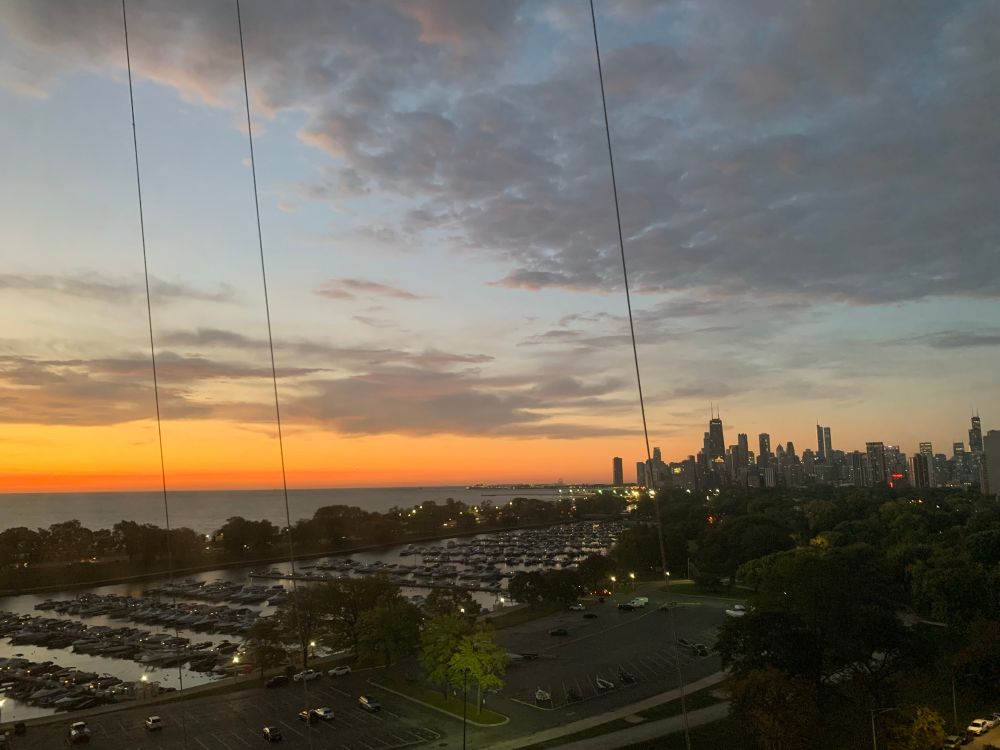 View of the Chicago skyline at sunrise, with Lake Michigan glowing orange at the horizon and dark clouds gathering above. The city’s skyscrapers are silhouetted on the right, while marinas and parking lots stretch across the foreground.