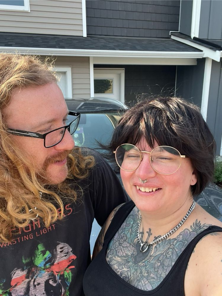 bailey and their partner standing in front of a house. bailey has a weird smile with their eyes closed. corwin is looking over at bailey and kinda beaming. they’re both wearing their forever collars haha. 