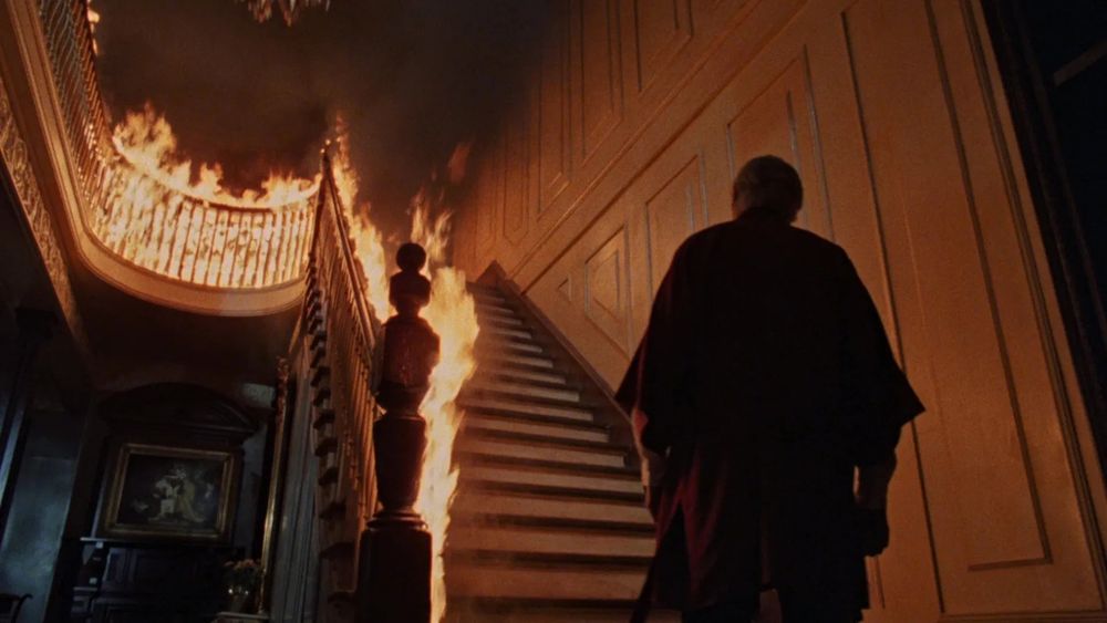 A still from the 1980 movie THE CHANGELING: George C. Scott stands at the foot of an elaborate Victorian staircase that is on fire. 