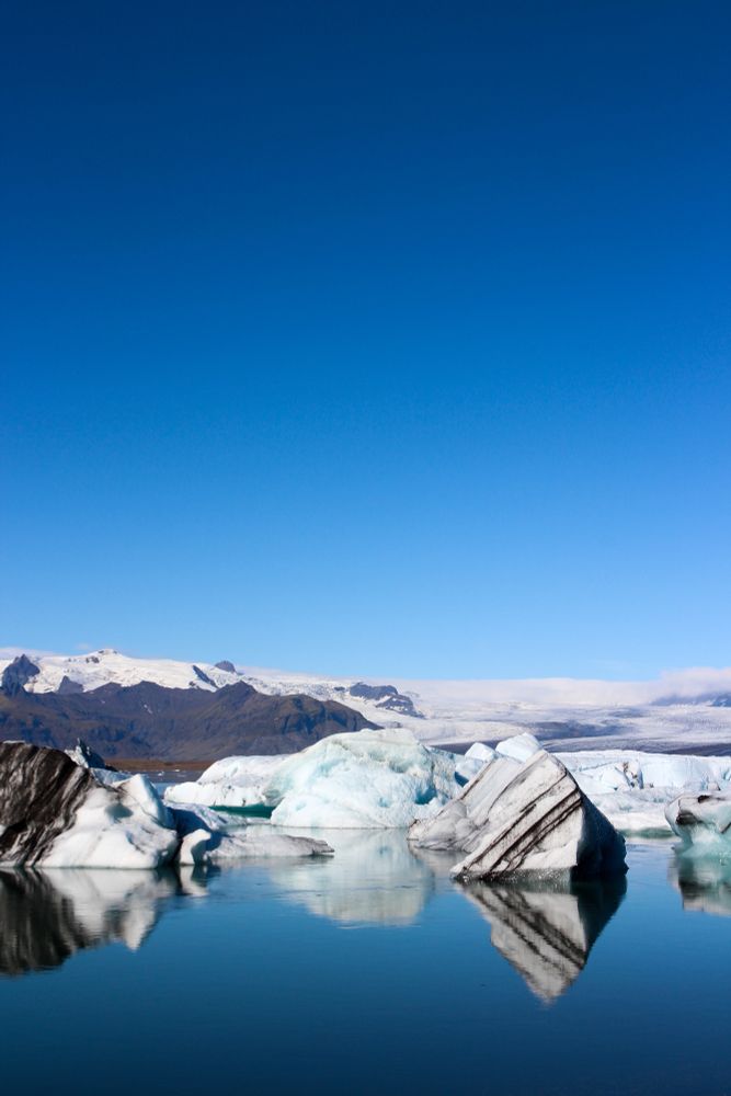 Photo du lac Jökulsárlón en Islande : on y voit un ciel très bleu sans nuage et des morceaux d'icebergs flottants sur une eau bleu unie. Derrière les blocs de glace se trouve une montagne avec de la neige sur son chapeau. 