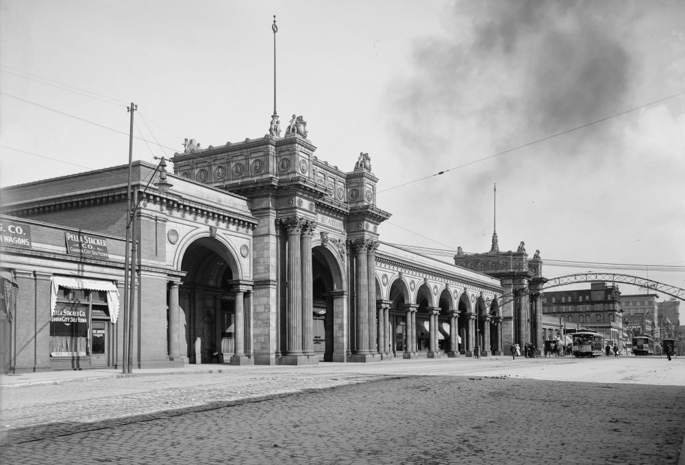A black and white photograph of the front facade of Columbus Union Station. The entrance features highly ornate decoration with large pillars and archways. A streetcar is visible on the street in front of the station.