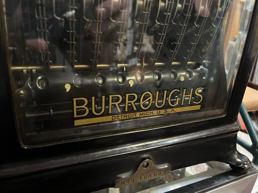 Photo of nameplate of Burroughs adding machine. Lettering is gold paint on glass, elegant serif'd copperplate capitals, with a rectangular block underneath with "DETROIT MICH. U. S. A.". The gold text is outlined in black as customary. The remarkable feature of the logo is that the O in "BURROUGHS" has been slashed. The machine is clearly very heavy and complex. Behind the glass is a register of a value in pennies: 0,000,000.00, with the commas and decimal point painted in gold also. The front of the machine has a flat unpainted metal face with an engraved serial number. 