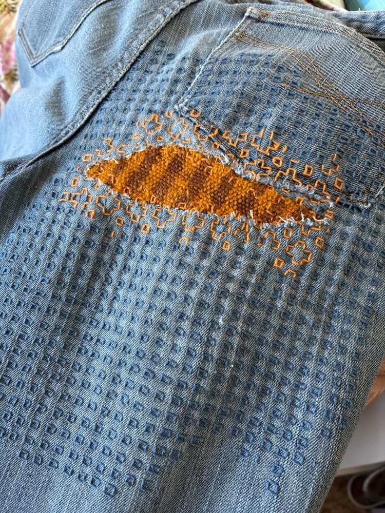 The back, right side of a pair of light blue jeans shows the pocket and the back thigh area that has been repaired. A large tear below the pocket is patched on the inside with a woven patch done in orange and brown. The patch has orange decorative stitching done around the edge and beyond for a short distance, and across the pocket and down the thigh has decorative stitching in blue. The blue stitching is all small squares aligned in a grid shape.