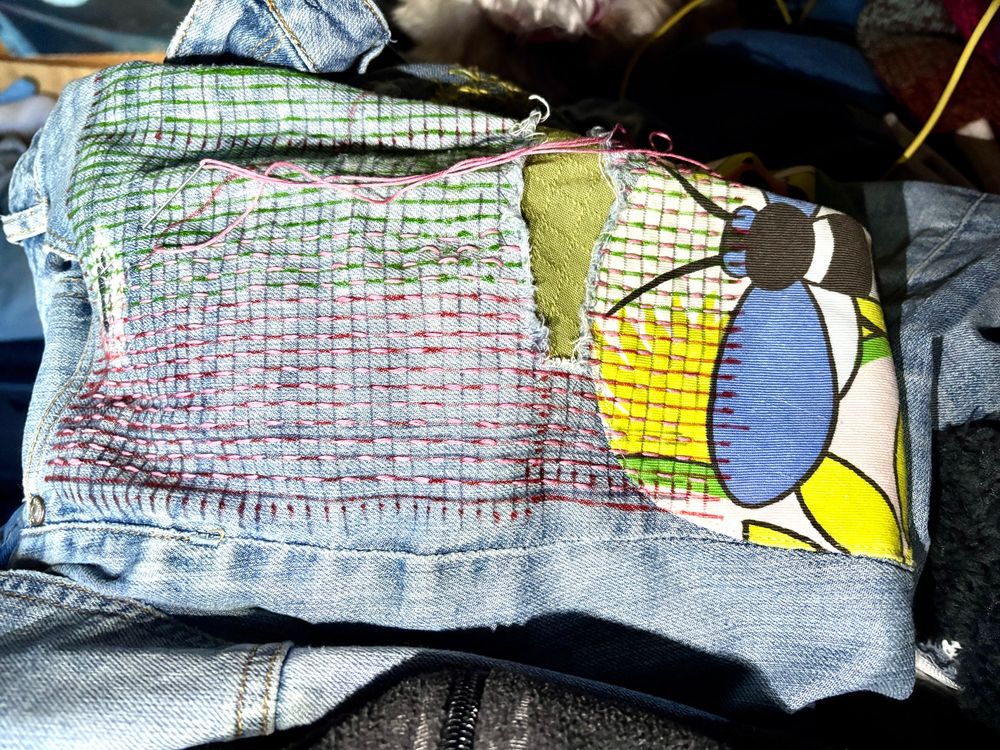 We see the upper thigh and waistband of a pair of pale blue jeans lying sideways across a lap that is fully obscured. The jeans have a 5mm grid drawn across them from the front pocket down to above the knee area. Some of the grid has pink running stitches across it that extend across a torn section that has a green piece of fabric stitched in place under the hole. On the right, covering the area above the knee of the jeans is a fabric patch sewn on top of brightly coloured vintage fabric featuring a garden scene; mostly we can see a bee with large blue wings. Some of the stitching and the drawn lines also cover the top of this patch just touching the bee’s wings.