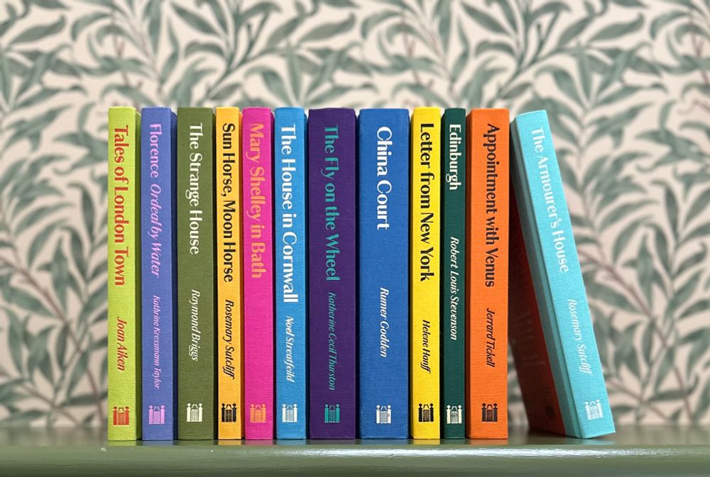 A shelf of colourful Manderley Press books spine out in front of William Morris willow wallpaper. 