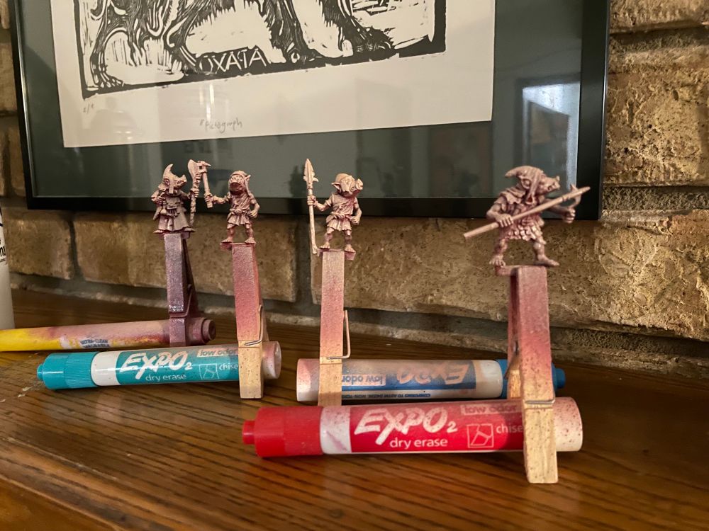Four 28mm orc miniatures that have been spray-primed attached to stands made out of clothespins and markers.