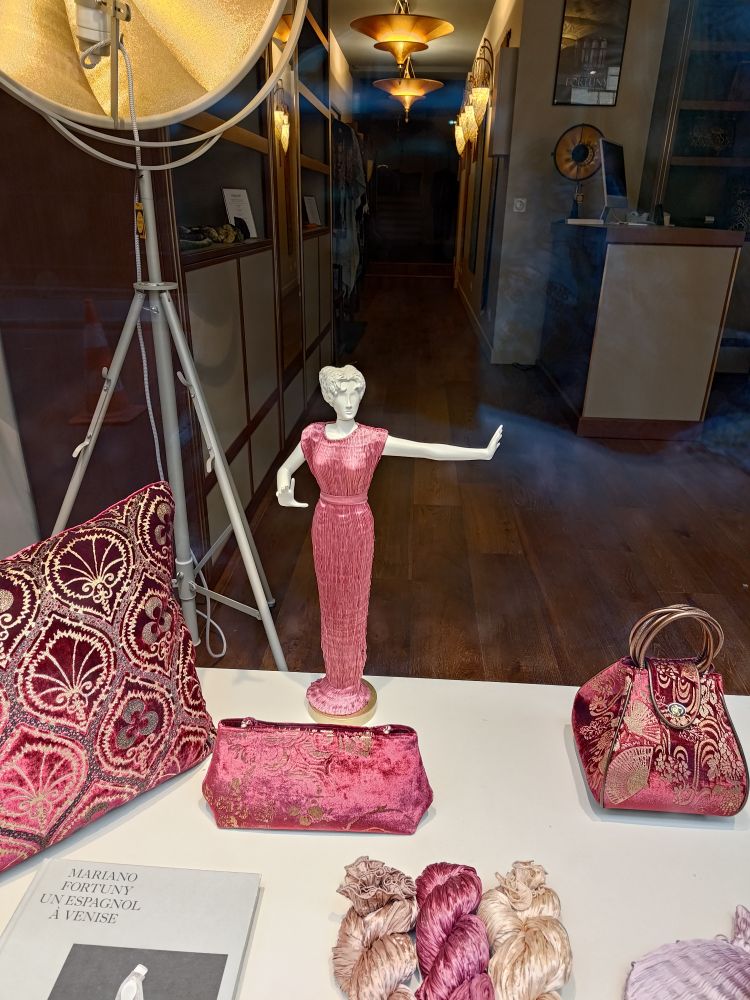 Photo de la vitrine de la boutique fortuny avec un coussin en velours rouge, des foulards en soie, un sac à main et une miniature de femme en robe delphos rouge