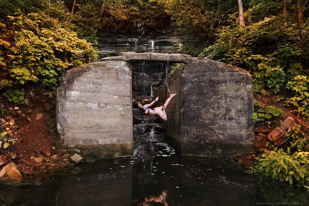 A woman tied in a TK, Futomomo and single ankle face down, suspended from a stone archway over water.