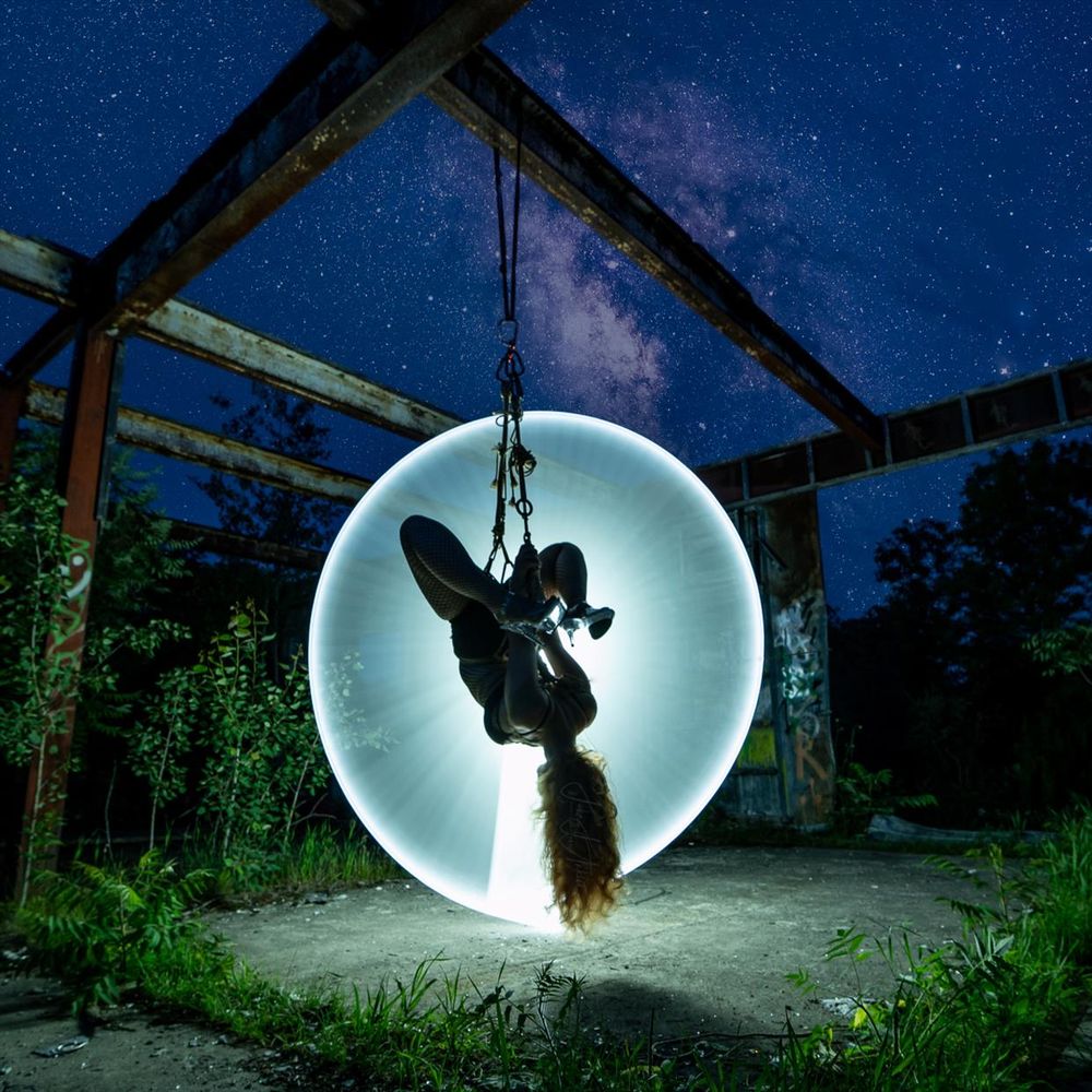 A woman tied in rope, suspended upside down from a steel beam in an abandoned building.  A white circle of light is behind her, illuminating her as a silhouette.