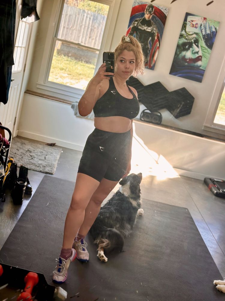 home gym mirror selfie in black sports bra and shorts. Keanu is laying behind Gabby, and Loki’s paws are in the edge of the pic.
