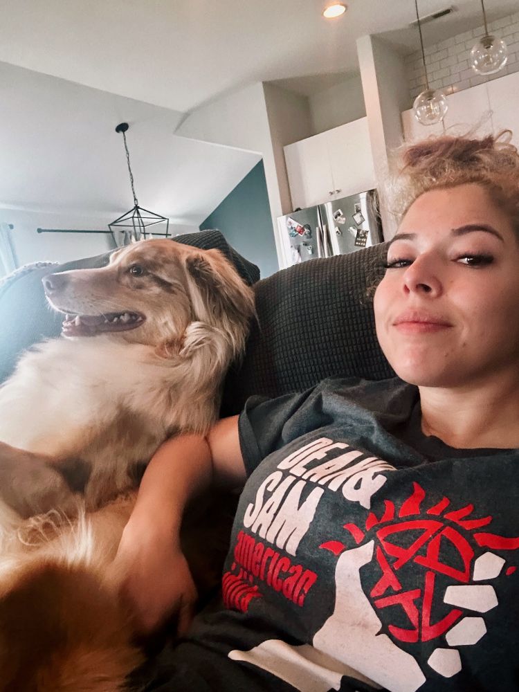 Gabby (wearing a Supernatural x American Idiot shirt) sitting with her Australian Shepherd Loki on the couch