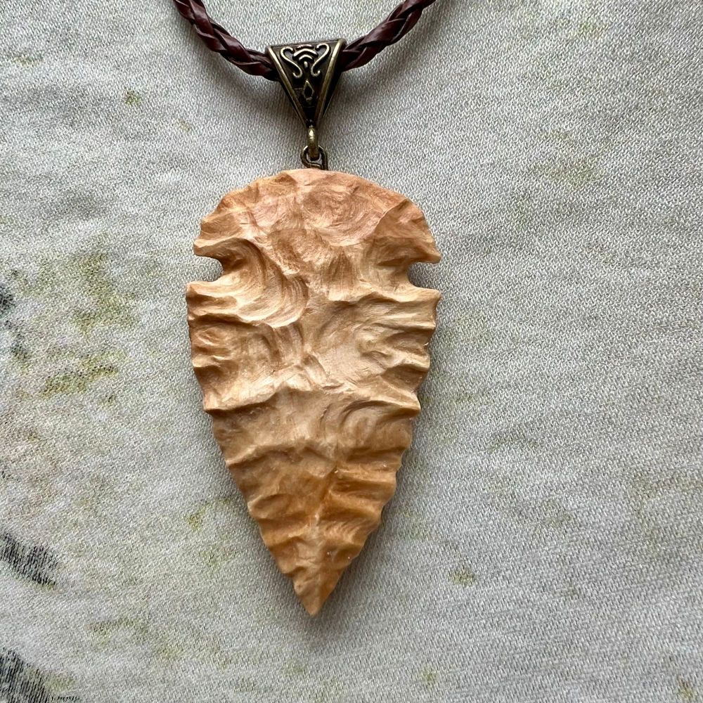 An arrowhead shape pendant, carved from light birch wood in such a way that the wooden texture mimics the texture of flaked stone, as if this wooden arrowhead was in fact knapped from stone like how the ancient warriors used to make them.