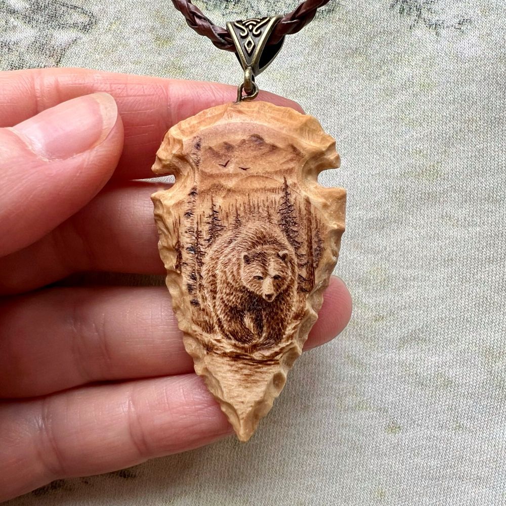 An arrowhead shape pendant, carved from wood in such a way that the wooden texture mimics the texture of flaked stone, as if this wooden arrowhead was in fact knapped from stone. On the one side, I wood burned an intricate image of a grizzly bear in the forest, walking toward you, pausing at the creekside.