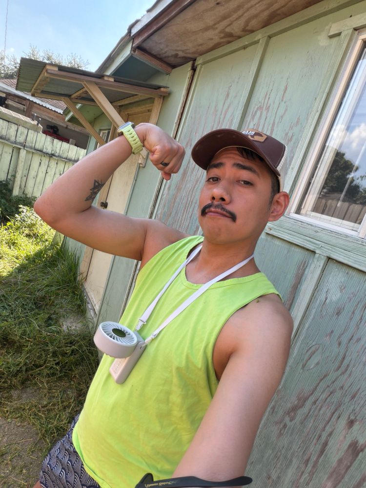 A Hispanic male, taking a selfie outdoors near a light green wooden building, wearing a brown cap, neon green sleeveless shirt, and white lanyard with a small white device. They are flexing one arm with a wristband and holding a blurred object. Background includes grass, a wooden fence, and part of another building.