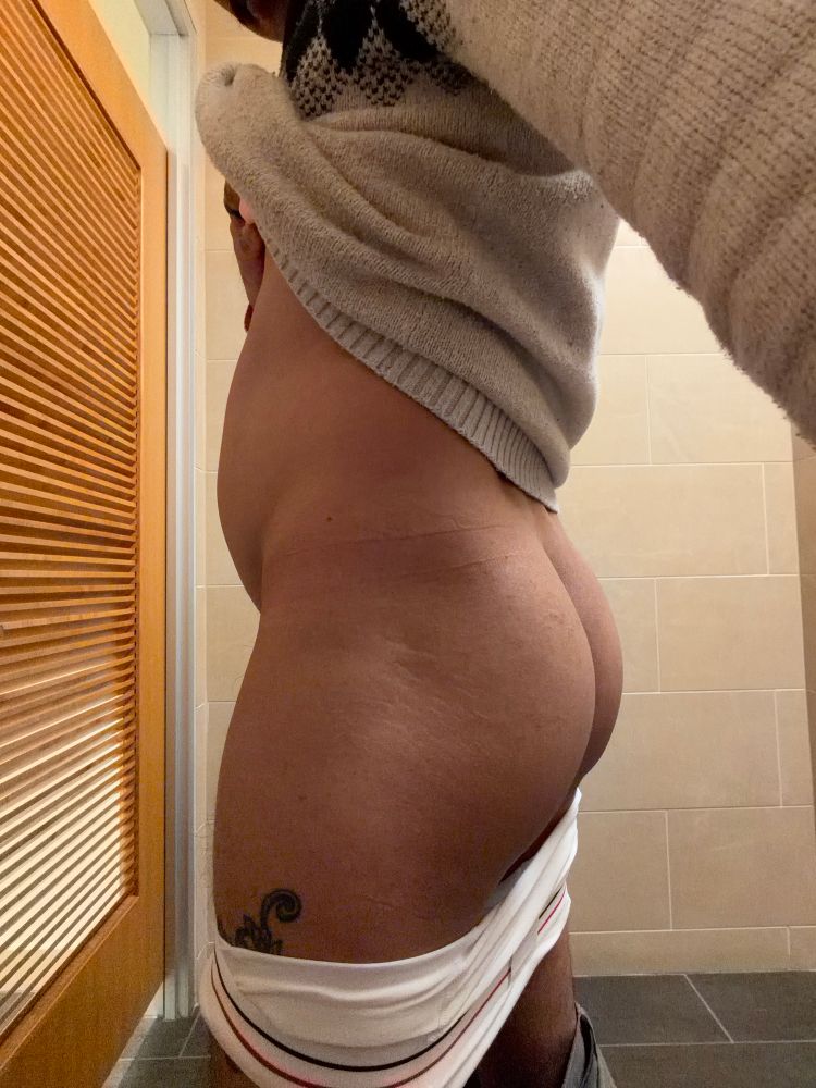 A male standing in profile as he pulls up his sweater, and his white boxer briefs are pulled down. 