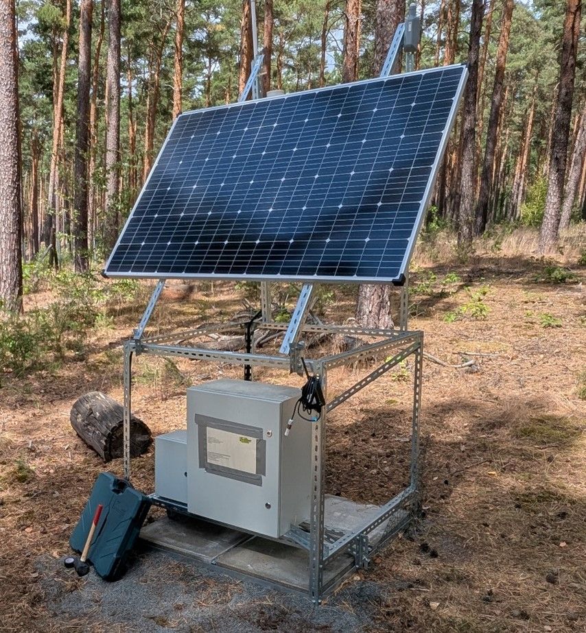 Solar-powered LoRaWan station with a large solar panel and control units mounted on a metal frame, installed in a forest clearing.