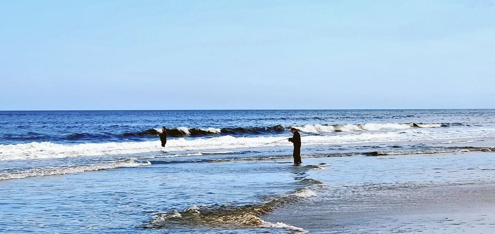 A beach at the danish north sea coast. Waves crash at the shore. The sky is blue. Two man with fishing rods standing in water, fishing.